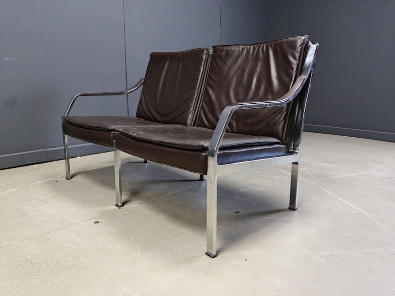 May include: A mid-century modern two-seater sofa with a dark brown leather seat and backrest. The sofa has a chrome-plated steel frame and legs. The design features a minimalist aesthetic, suitable for a living room or office space.