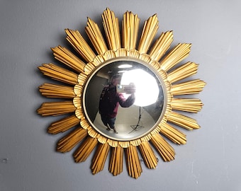 Puede incluir: Un espejo dorado con forma de sol con un centro redondo y reflectante. El marco del espejo presenta rayos puntiagudos y radiantes. El espejo está montado en una pared gris, reflejando la habitación y la persona que toma la foto.