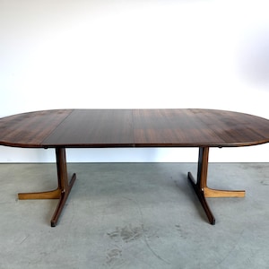 Peut inclure: Une table à manger ovale en bois avec une finition en grain de bois brun foncé. La table dispose d'une rallonge centrale et est soutenue par deux pieds en forme de T, chacun doté de deux pieds s'étendant vers l'extérieur pour la stabilité.
