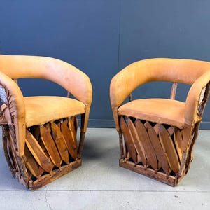 May include: A pair of armchairs featuring light tan leather upholstery on the curved backrests and seats. The base of each chair is constructed from interwoven wooden slats and natural branch-like supports, creating a distinct structural frame.