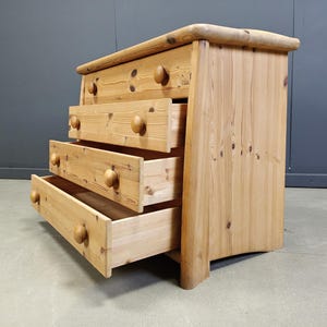 May include: A light-coloured wooden chest of drawers with four drawers, three of which are open. The chest has rounded wooden knobs and a curved top. The wood grain is visible throughout, giving it a natural look.