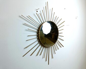 Puede incluir: Un espejo de sol dorado con una superficie redonda y reflectante. El espejo tiene un diseño de estrella con muchos rayos finos y puntiagudos.