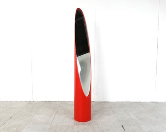 May include: A tall, red, cylindrical mirror with a curved, oval-shaped reflective surface.