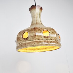 May include: A ceramic pendant light fixture with a brown and white glaze. The light has a round, bell-shaped shade with multiple circular cutouts. The light is turned on and emits a warm yellow glow.