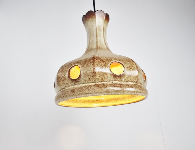 May include: A ceramic pendant light fixture with a brown and white glaze. The light has a round, bell-shaped shade with multiple circular cutouts. The light is turned on and emits a warm yellow glow.