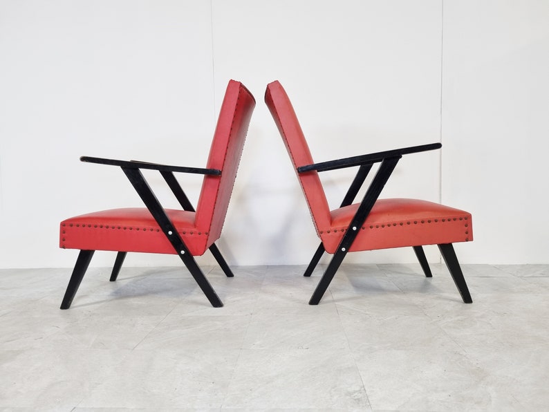May include: Two red armchairs with black wooden frames. The chairs have a simple, mid-century modern design.