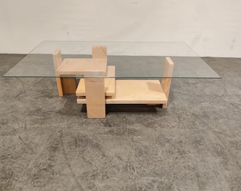 May include: A modern coffee table with a glass top and a wooden base. The base is made of multiple stacked pieces of wood, creating a geometric design.