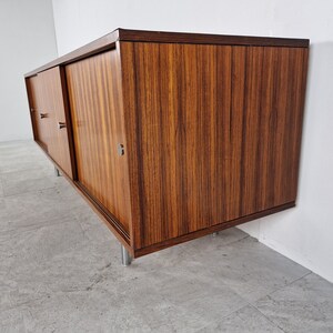 May include: A mid-century modern sideboard with sliding doors made of dark wood with a wood grain pattern. The sideboard has metal legs and pulls.