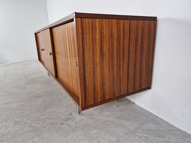 May include: A mid-century modern sideboard with sliding doors made of dark wood with a wood grain pattern. The sideboard has metal legs and pulls.