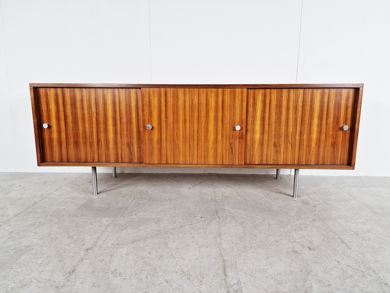 May include: A mid-century modern sideboard with three sliding doors and metal legs. The cabinet is made of wood with a rich brown finish and has a sleek, minimalist design.