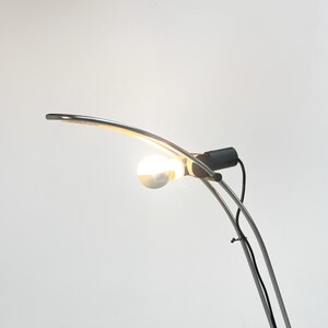 Pode incluir: Um candeeiro de p&eacute; de arco prateado com uma l&acirc;mpada branca. O candeeiro tem um cabo preto e um interruptor preto.