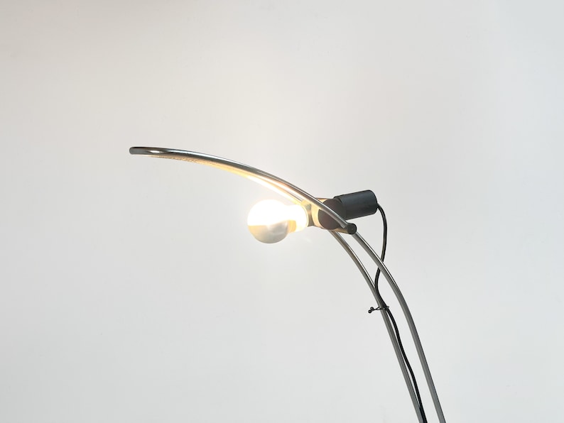 Pode incluir: Um candeeiro de p&eacute; de arco prateado com uma l&acirc;mpada branca. O candeeiro tem um cabo preto e um interruptor preto.