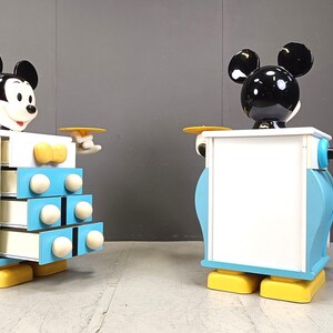 Peut inclure: Deux figurines de Mickey Mouse, chacune avec une tête et des oreilles noires, un visage blanc et un corps bleu et blanc. La figurine de gauche a un plateau dans sa main et un ensemble de tiroirs dans son corps. La figurine de droite a un plateau dans sa main et un panneau blanc dans son corps.