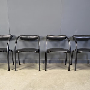 Peut inclure: Quatre chaises noires avec accoudoirs et dossiers incurvés. Les chaises ont un design minimaliste avec des cadres en métal noir et des assises en cuir noir. Les chaises sont disposées en rangée contre un mur gris.