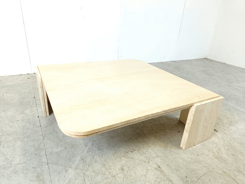 May include: A square, light-colored wood coffee table with rounded edges and a unique, layered base.