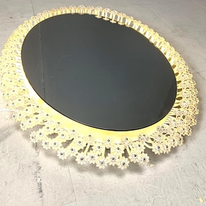 Op de afbeelding: Een ronde spiegel met een decoratief gouden frame. Het frame is versierd met kleine, witte, bloemvormige details.
