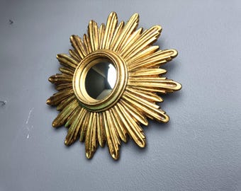 Puede incluir: Un espejo dorado con forma de sol, con un centro redondo y reflectante. El espejo tiene un diseño detallado y texturizado con rayos radiantes. Está montado en una pared gris, creando un contraste llamativo. Una pieza decorativa para el hogar.