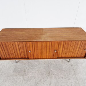 May include: A mid-century modern wooden cabinet with three sliding doors and metal handles. The cabinet is made of a light brown wood with a wood grain pattern. The cabinet is sitting on four metal legs.