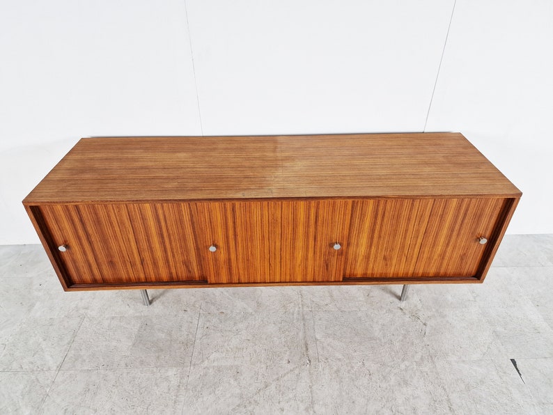 May include: A mid-century modern wooden cabinet with three sliding doors and metal handles. The cabinet is made of a light brown wood with a wood grain pattern. The cabinet is sitting on four metal legs.