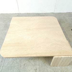 May include: A square, light beige travertine coffee table with a rounded edge and a unique, stacked base.