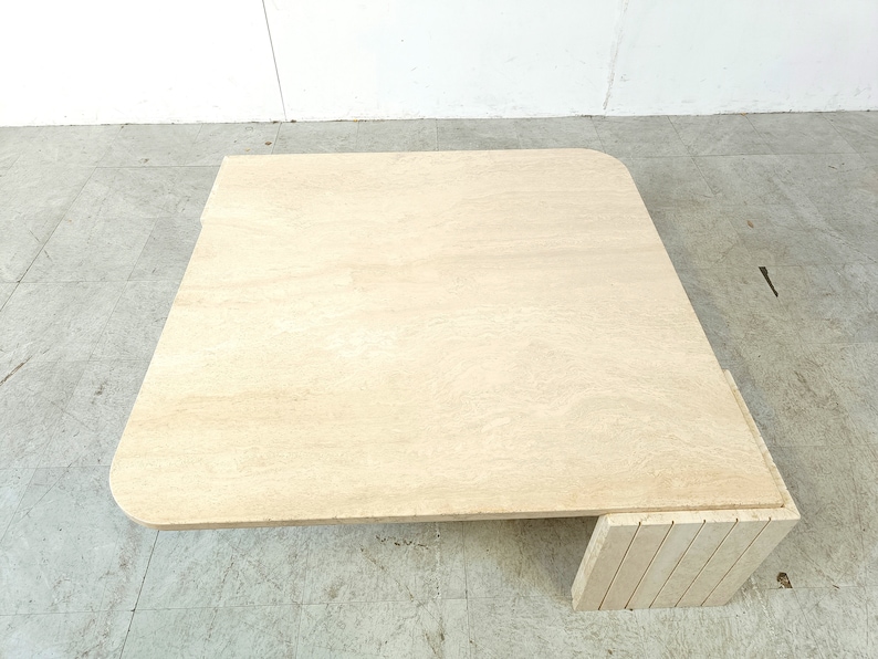 May include: A square, light beige travertine coffee table with a rounded edge and a unique, stacked base.