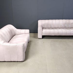 May include: Two light pink sofas with a modern design. One sofa has a curved back and the other has a straight back. Both sofas have a ribbed texture and are set against a dark grey wall.