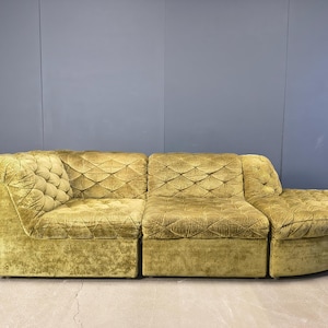 May include: An olive green vintage sectional sofa with a corduroy texture. The sofa has a tufted corner piece, two central sections with diamond stitching, and a rounded ottoman. The sofa is set against a grey-blue wall.