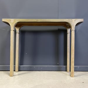 May include: A light-coloured, rectangular console table with a curved design. The table has four vertical legs and a horizontal top. The legs and top are made of a light-coloured material, possibly bamboo or rattan, with a ribbed texture.