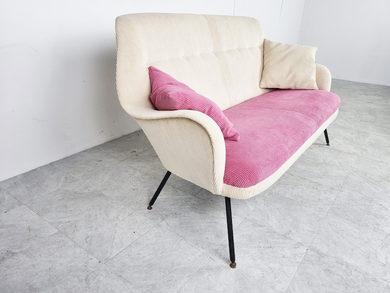 May include: A white and pink fabric sofa with black metal legs. The sofa has a tufted back and a pink cushion.