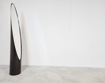 May include: A tall, black, free-standing mirror with an oval-shaped reflective surface. The mirror is on a white tiled floor.