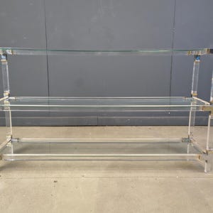 May include: A clear acrylic and metal shelf unit with two glass shelves. The frame features clear acrylic supports and metal accents. The unit is designed for display or storage and is suitable for various home decor styles.