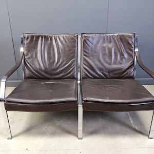 May include: A two-seat brown leather sofa with a chrome frame. The cushions are a dark brown color, and the arms are a darker shade of brown. The sofa has a modern, minimalist design.