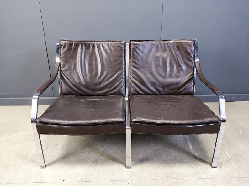 May include: A two-seat brown leather sofa with a chrome frame. The cushions are a dark brown color, and the arms are a darker shade of brown. The sofa has a modern, minimalist design.