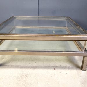 May include: A square coffee table with a glass top and a lower glass shelf. The frame is made of a brushed metal material. The table is approximately 122 cm long, 122 cm wide, and 41 cm high.