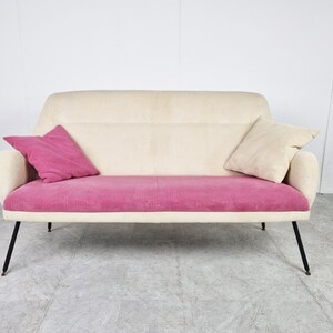 May include: A white and pink two-seater sofa with black metal legs. The sofa has a tufted back and a corduroy seat.