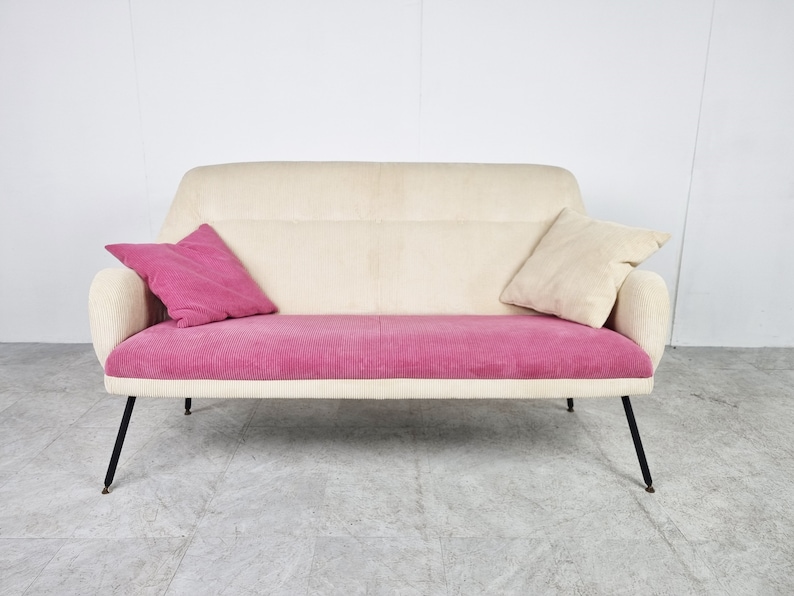 May include: A white and pink two-seater sofa with black metal legs. The sofa has a tufted back and a corduroy seat.