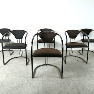 Peut inclure: Ensemble de six chaises de salle à manger en métal noir avec sièges en tissu marron. Les chaises ont un dossier et des accoudoirs incurvés.