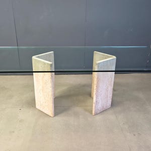 Op de afbeelding: Een rechthoekige glazen tafel met een helder blad en twee travertijnen stenen steunen. De steunen zijn lichtbeige en hebben een V-vormig ontwerp. De tafel heeft een modern design en staat op een betonnen vloer.