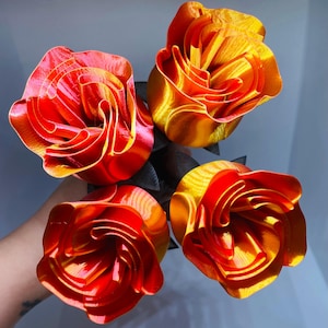 May include: A bouquet of four vibrant, layered roses. The petals transition from deep red to bright orange and yellow, with a glossy finish. The stems are black, creating a striking contrast. A unique floral arrangement.