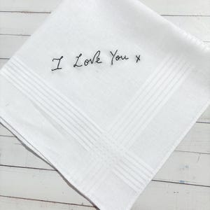 May include: A white square handkerchief featuring a subtle woven striped border pattern and the phrase I Love You x embroidered in black thread in the centre. The fabric has a smooth texture and is folded into a diamond orientation.