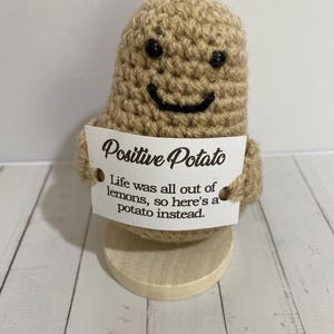 May include: A handmade crocheted potato figure with a smiling face and black button eyes. It holds a sign that says "Positive Potato" and "Life was all out of lemons, so here's a potato instead."