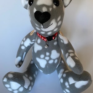 May include: A grey and white paw print patterned teddy bear with black eyes and a red collar with white flowers and a silver charm.