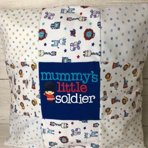May include: A white and blue patchwork pillow with a blue fabric panel in the centre that reads "mummy's little soldier". The pillow is decorated with a variety of patterns, including stars, animals, and a red and blue soldier.