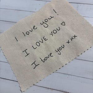 May include: A beige fabric square with the handwritten phrase "I love you" repeated three times in black ink. The fabric has a raw, unfinished edge and is set against a white wooden background.