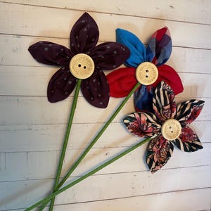 May include: Three fabric flowers with wooden button centres and green stems. The flower petals are made from patterned fabrics in burgundy, red, blue, and black. The wooden buttons have the text "Handmade with love" around the edges.