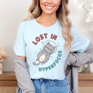 May include: A light blue t-shirt with a cartoon cat wearing a space helmet. The text "LOST IN HYPERFOCUS" is printed on the shirt.