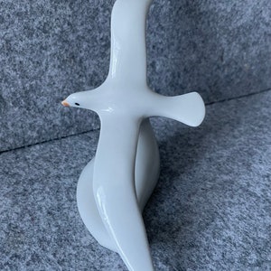 May include: White porcelain figurine of a seagull in flight. The seagull is depicted with its wings spread wide and its head tilted slightly to the side. The figurine is a simple and elegant design.