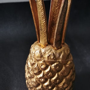 May include: A gold pineapple-shaped toothpick holder with a textured surface. The holder has a set of toothpicks sticking out of the top.