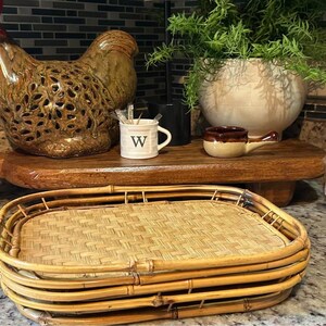 May include: A decorative rooster figurine with a brown finish and openwork design sits on a wooden tray. A small white mug with a black "W" and a small brown and beige handled dish are also on the tray. A woven bamboo tray is in the foreground.