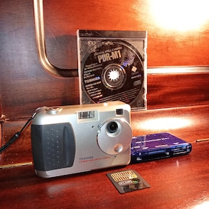 Puede incluir: Una cámara digital plateada Toshiba con un agarre negro, un CD-ROM, un lector de tarjetas azul y una tarjeta de memoria se muestran sobre una superficie de madera. La cámara tiene el texto "TOSHIBA 1.5 MEGA PIXEL DIGITAL CAMERA".
